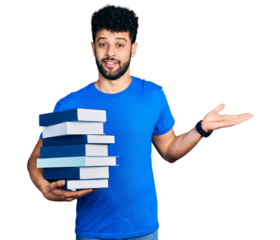 Young arab man with beard holding a pile of books celebrating achievement with happy smile and winner expression with raised hand