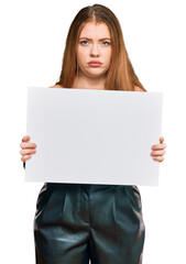 Young beautiful redhead woman holding blank empty banner thinking attitude and sober expression looking self confident