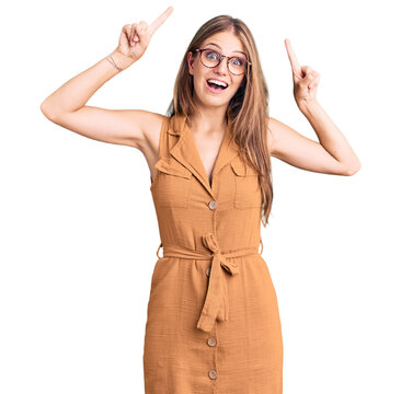 Young beautiful blonde woman wearing casual clothes and glasses smiling amazed and surprised and pointing up with fingers and raised arms.