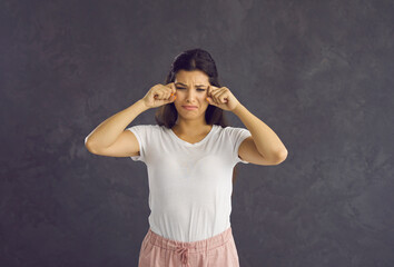 Capricious and dissatisfied young woman with frowning face cries and wipes away tears. Upset and unhappy Caucasian woman who did not get what she wanted. Isolated on gray background.