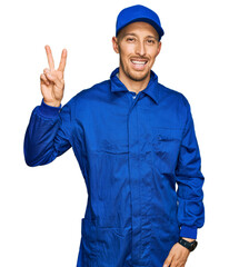 Bald man with beard wearing builder jumpsuit uniform smiling looking to the camera showing fingers doing victory sign. number two.
