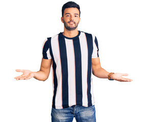 Naklejka premium Young handsome hispanic man wearing striped tshirt clueless and confused expression with arms and hands raised. doubt concept.