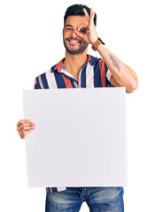 Young handsome hispanic man holding cardboard banner with blank space smiling happy doing ok sign with hand on eye looking through fingers