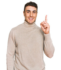 Fototapeta premium Hispanic young man wearing casual turtleneck sweater pointing finger up with successful idea. exited and happy. number one.