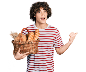 Caucasian teenager holding wicker basket with bread pointing thumb up to the side smiling happy with open mouth
