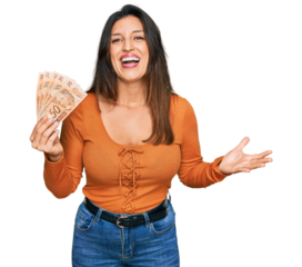 Beautiful hispanic woman holding 50 brazilian real banknotes celebrating victory with happy smile and winner expression with raised hands