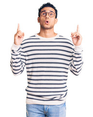 Naklejka premium Hispanic handsome young man wearing casual clothes and glasses amazed and surprised looking up and pointing with fingers and raised arms.