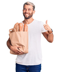 Young handsome blond man holding paper bag with bread smiling happy and positive, thumb up doing...