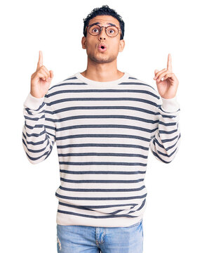 Hispanic handsome young man wearing casual clothes and glasses amazed and surprised looking up and pointing with fingers and raised arms.