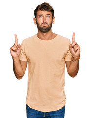 Handsome young man with beard wearing casual tshirt pointing up looking sad and upset, indicating...
