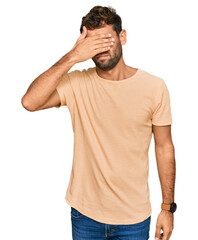 Fototapeta premium Handsome young man with beard wearing casual tshirt covering eyes with hand, looking serious and sad. sightless, hiding and rejection concept