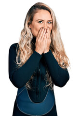 Young blonde woman wearing diver neoprene uniform laughing and embarrassed giggle covering mouth...