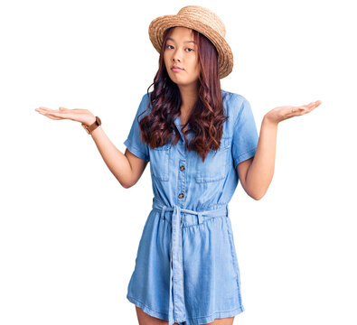 Young beautiful chinese girl wearing summer hat clueless and confused expression with arms and hands raised. doubt concept.