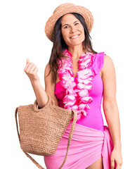 Young beautiful brunette woman wearing swimwear and hawaiian lei holding bag looking positive and...