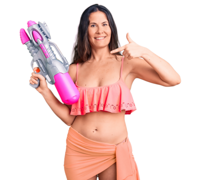 Young beautiful brunette woman wearing bikini holding water gun pointing finger to one self smiling happy and proud