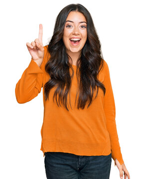 Beautiful brunette young woman wearing casual orange sweater pointing finger up with successful idea. exited and happy. number one.