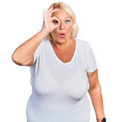 Middle age blonde woman wearing casual white t shirt doing ok gesture shocked with surprised face, eye looking through fingers. unbelieving expression.