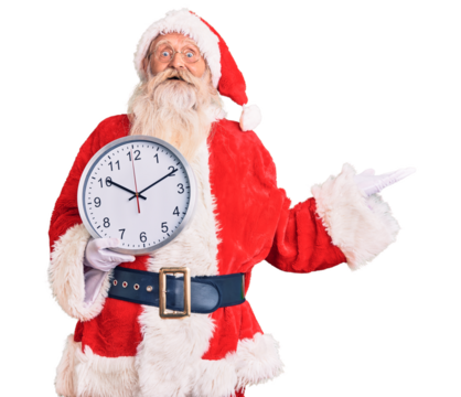 Old senior man with grey hair and long beard wearing santa claus costume holding clock celebrating victory with happy smile and winner expression with raised hands