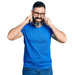 Hispanic man with beard wearing casual t shirt and glasses covering ears with fingers with annoyed expression for the noise of loud music. deaf concept.