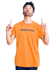 Young hispanic man wearing t shirt with happiness word message amazed and surprised looking up and pointing with fingers and raised arms.