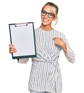Beautiful blonde business woman holding clipboard with blank space pointing finger to one self smiling happy and proud