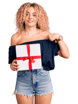 Young blonde woman with curly hair holding gift pointing finger to one self smiling happy and proud