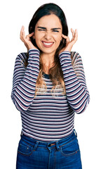 Obraz na płótnie Canvas Beautiful hispanic woman wearing casual striped shirt covering ears with fingers with annoyed expression for the noise of loud music. deaf concept.