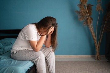 Depressed young woman suffering migraine sitting alone on the bed with hands on head feel stress,...