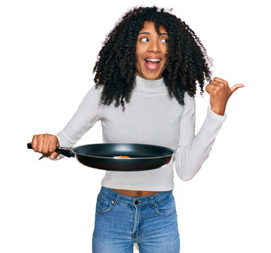 Young african american girl holding skillet with fried egg pointing thumb up to the side smiling happy with open mouth