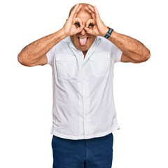 Fototapeta premium Mature middle east man with mustache wearing casual white shirt doing ok gesture like binoculars sticking tongue out, eyes looking through fingers. crazy expression.