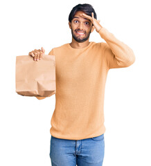 Fototapeta premium Handsome hispanic man holding take away paper bag stressed and frustrated with hand on head, surprised and angry face