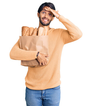 Handsome hispanic man holding paper bag with bread stressed and frustrated with hand on head, surprised and angry face