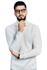 Young arab man with beard wearing elegant turtleneck sweater and glasses with hand on chin thinking about question, pensive expression. smiling and thoughtful face. doubt concept.