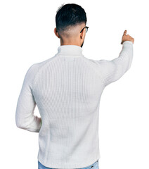 Young arab man with beard wearing elegant turtleneck sweater and glasses posing backwards pointing ahead with finger hand