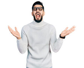 Fototapeta premium Young arab man with beard wearing elegant turtleneck sweater and glasses celebrating mad and crazy for success with arms raised and closed eyes screaming excited. winner concept
