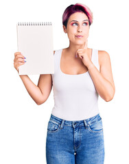 Young beautiful woman with pink hair holding notebook serious face thinking about question with...