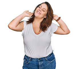 Obraz na płótnie Canvas Young plus size woman wearing casual white t shirt relaxing and stretching, arms and hands behind head and neck smiling happy