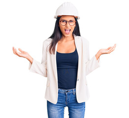 Young beautiful latin girl wearing architect hardhat and glasses crazy and mad shouting and yelling with aggressive expression and arms raised. frustration concept.