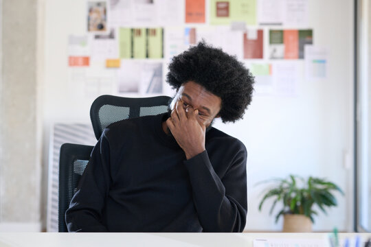 Black businessman with afro look sitting at his desk in the office. He rubs his eyes because he is tired. Overtime at work