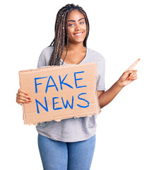 Young african american woman with braids holding fake news banner smiling happy pointing with hand...