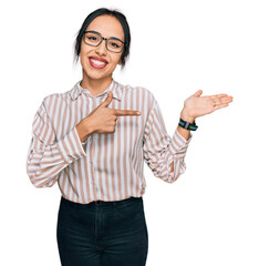 Young hispanic girl wearing casual clothes and glasses amazed and smiling to the camera while presenting with hand and pointing with finger.