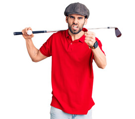 Young handsome man with beard playing golf holding club and ball annoyed and frustrated shouting...