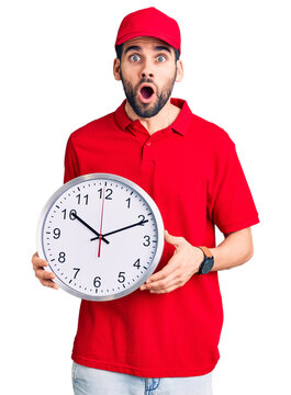 Young handsome man with beard wearing delivery uniform holding big clock scared and amazed with open mouth for surprise, disbelief face