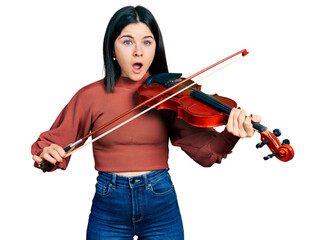 Young brunette woman with blue eyes playing violin afraid and shocked with surprise and amazed expression, fear and excited face.