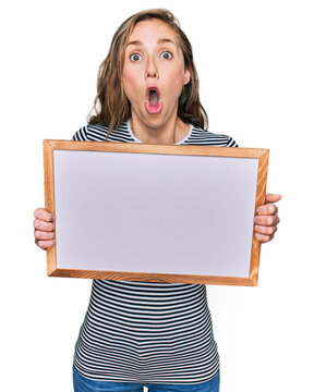 Young blonde woman holding empty white board scared and amazed with open mouth for surprise, disbelief face