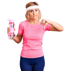 Young blonde woman wearing sportswear holding water bottle with angry face, negative sign showing...