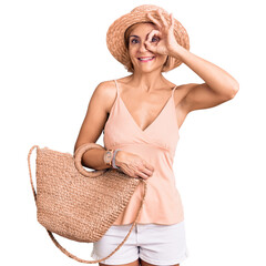 Young blonde woman wearing summer hat holding bag smiling happy doing ok sign with hand on eye...