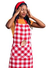 Young indian girl wearing professional baker apron with hand on head for pain in head because stress. suffering migraine.