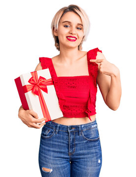 Young beautiful blonde woman holding gift pointing finger to one self smiling happy and proud