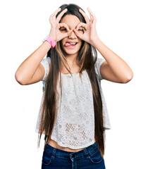 Obraz na płótnie Canvas Young brunette girl with long hair wearing white shirt doing ok gesture like binoculars sticking tongue out, eyes looking through fingers. crazy expression.